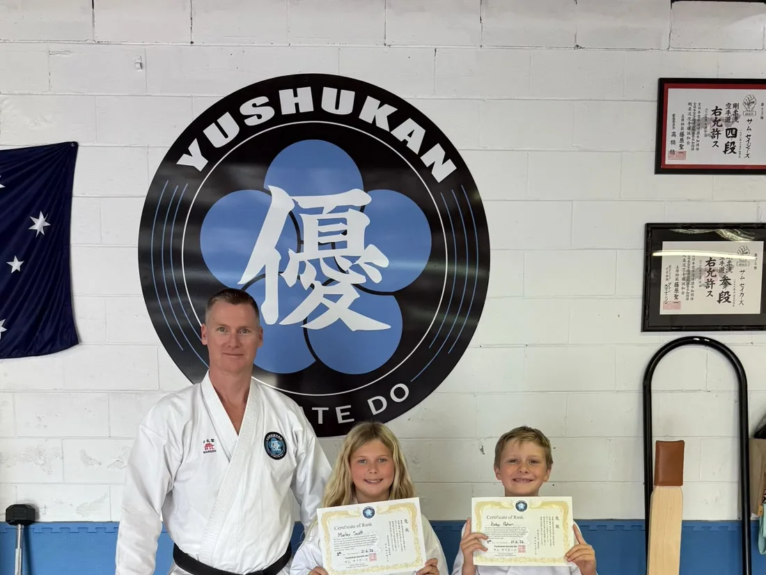 Sensei Sam Seigers with two students holding grading certificates at Yushukan Karate, dan certificates visible on the wall