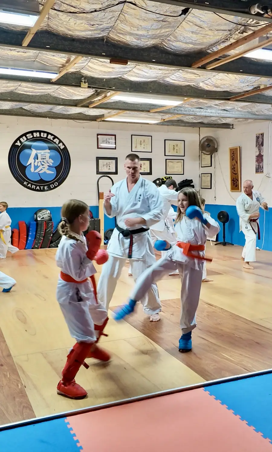 Two Yushukan Karate students sparring in the Honbu Dojo — controlled kumite with protective gear, supervised by Sensei Sam